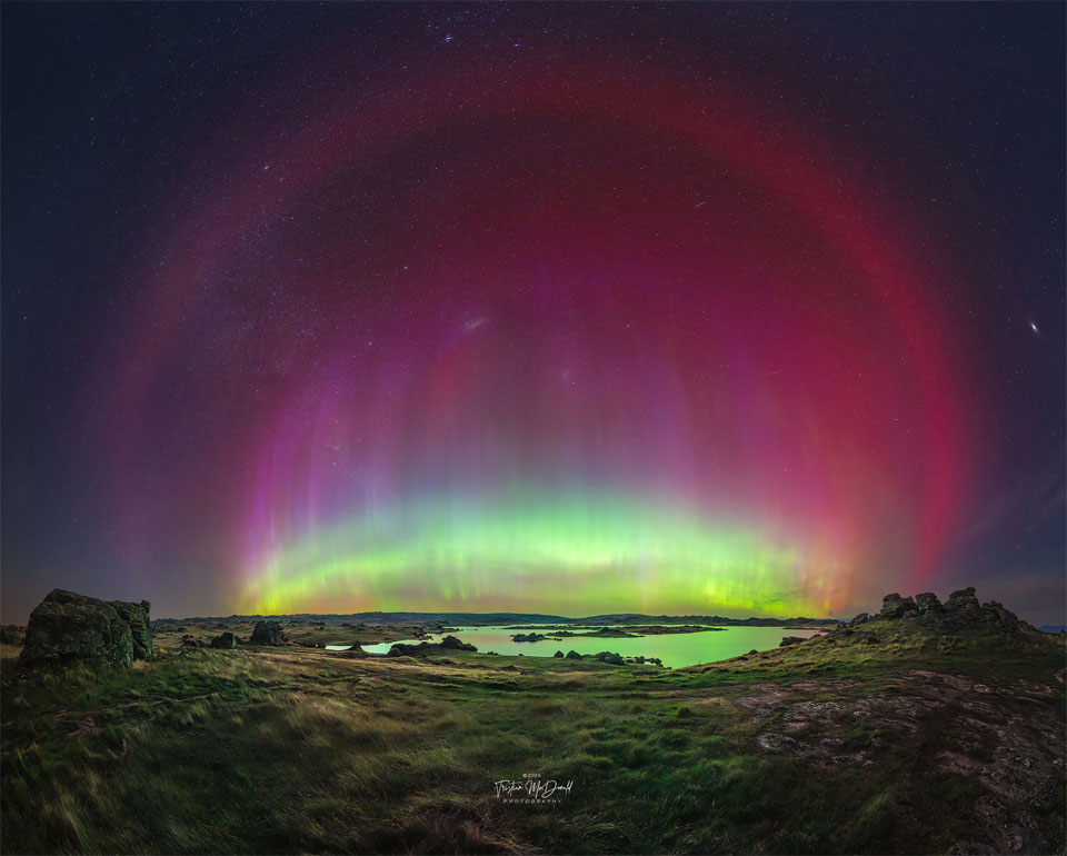 A flat landscape with a pond is imaged at night below
a starfield. A multicolored aurora is seen in an arc across
 the image center. Around this arc is another red arc that
is particularly smooth. 
Please see the explanation for more detailed information.