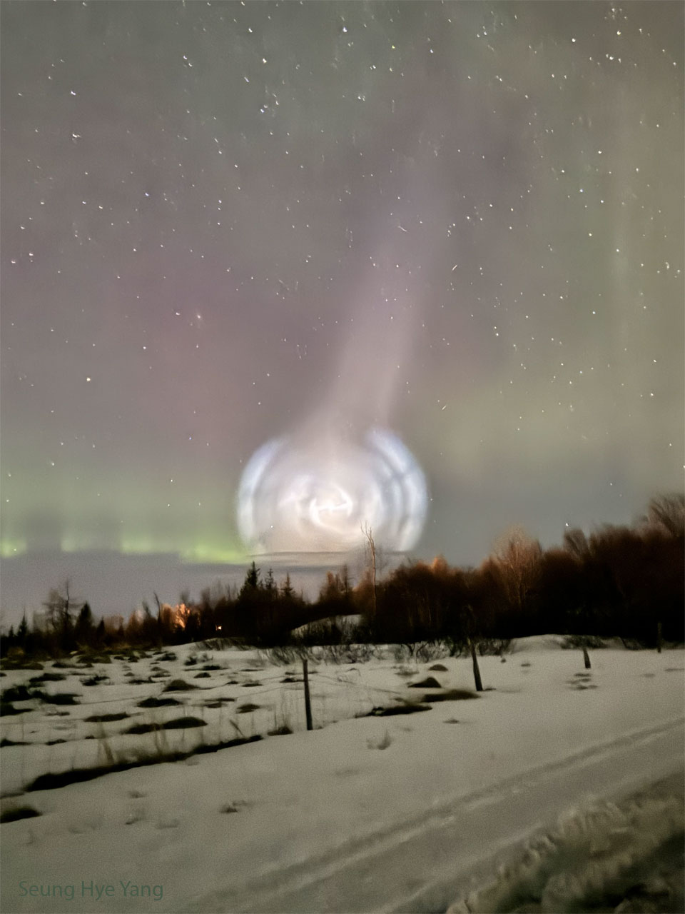 A field of snow is shown, lined with trees along the back.
Above the horizon is an unusual white spiral cloud. 
Stars dot the background, and faint green and red aurora
are also visible. 
Please see the explanation for more detailed information.