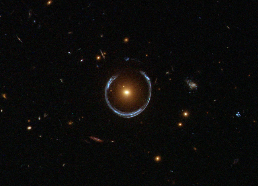 A dark star field with several small galaxies 
features a yellowish galaxy in the center. Surrounding
this galaxy is a blue nearly circular horseshoe shaped
object. 
Please see the explanation for more detailed information.