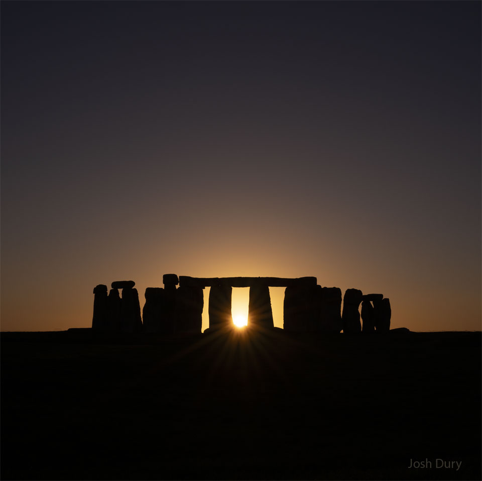The sun is setting across a field in a clear sky.
In the field are the famous stones of Stonehenge. The
Sun peaks out from the center of the stone array.
Please see the explanation for more detailed information.