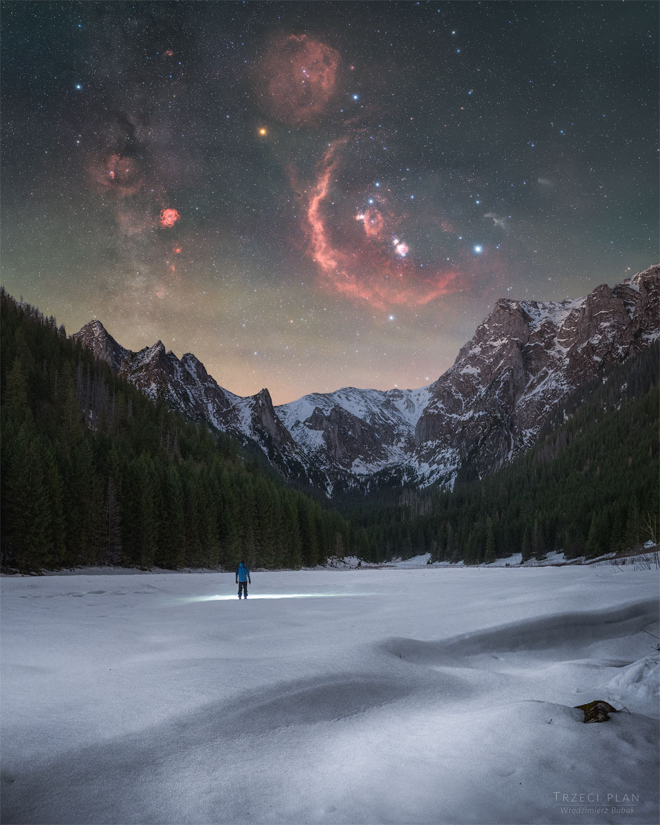 A person stands in the distance on a snowy landscape shining
a flashlight on the ground. Trees and snow capped mountains are
on the horizon. In the starry sky above, several iconic red
nebulas are visible. 
Please see the explanation for more detailed information.