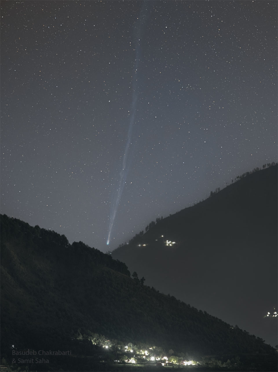 A starry sky is seen above two tree-covered mountains
that slope up on both sides of the image. In the central
valley the head of a comet is seen, with a long tail 
flowing nearly vertically upward toward the top of the 
tall frame. 
Please see the explanation for more detailed information.