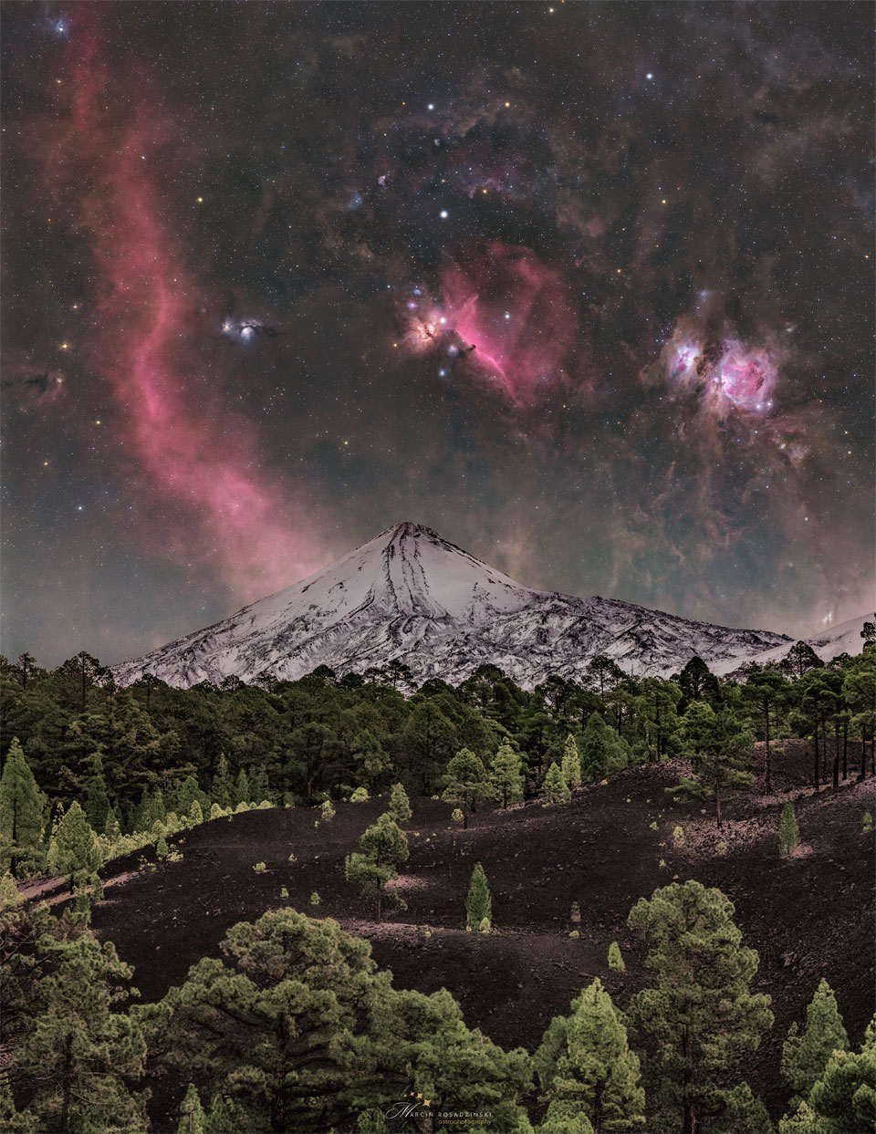 A starry sky is seen above a snowy volcano peak. The sky is
bright with many stars and red nebula. In the foreground is dark
soil and green evergreen trees. 
Please see the explanation for more detailed information.