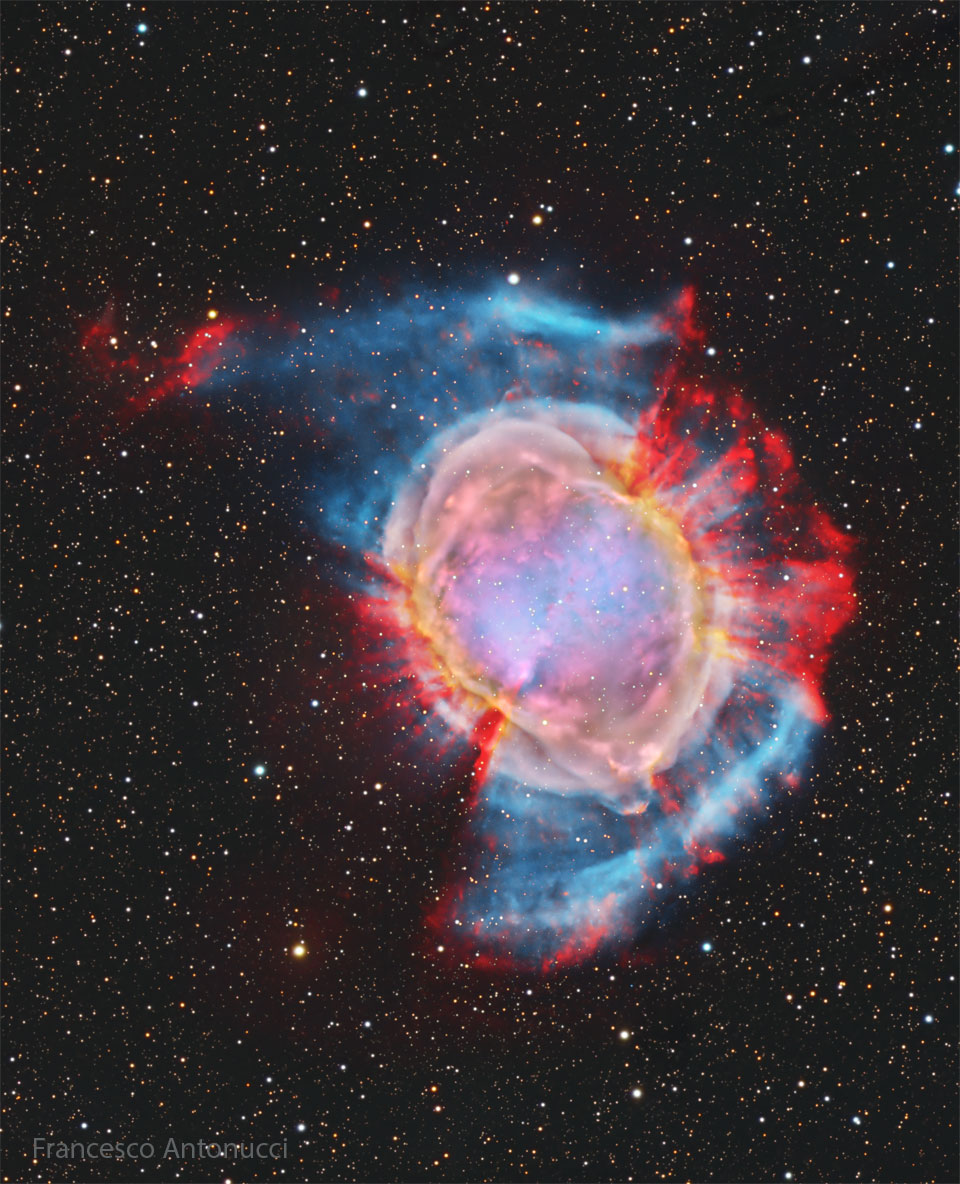 A starfield surrounds a large and colorful nebula.
Red spokes poke out from the outside of the nebula in
some directions, while a blue haze appears in other
directions. The nebula center is multicolored. 
Please see the explanation for more detailed information.